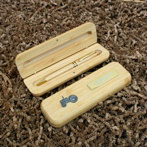 May include: A light-colored wooden pen and case set. The pen has gold-colored accents. The case is open, revealing the pen inside. The closed case has a small metal tractor emblem and a gold-colored nameplate.