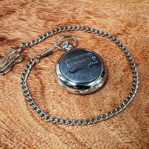Puede incluir: Un reloj de bolsillo plateado con una cadena. La esfera del reloj tiene un fondo negro con un autobús de dos pisos plateado grabado en él.