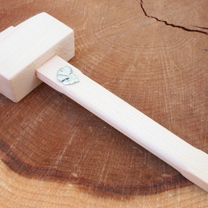 Puede incluir: Un mazo de madera con un acabado marrón claro y un pequeño amuleto de metal en el mango. El mazo está sobre una superficie de madera.