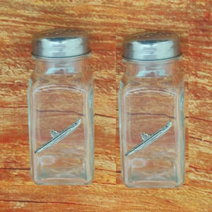 May include: A set of two glass salt and pepper shakers with silver lids. Each shaker features a small pewter submarine design.