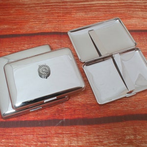 Op de afbeelding: Meerdere glanzende zilverkleurige metalen sigarettenkokers. Eén koker is open en toont de binnenkant. De andere zijn gesloten, met een decoratief cirkelvormig embleem erop. De kokers zijn rechthoekig met afgeronde hoeken.