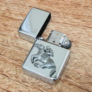 May include: A silver lighter with a raised design of a person riding a horse. The lighter is open and the lid is laying next to it.