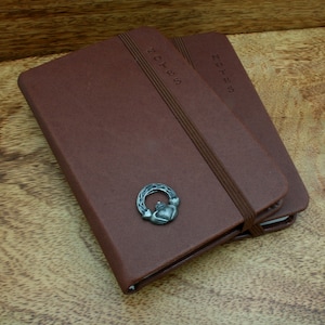May include: Two brown leather-bound notebooks with the word "NOTES" embossed on the cover. The top notebook has a silver Celtic knot charm attached.