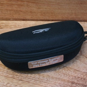 Könnte beinhalten: Schwarze Reißverschlusstasche für Sonnenbrillen mit einem silbernen Metallplättchen mit der Aufschrift "Ihre Botschaft hier kostenlos graviert".