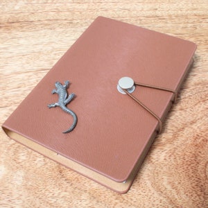 May include: A brown leather journal with a silver lizard charm and an elastic closure. The journal is closed and has a smooth texture. The lizard charm is detailed and adds a decorative element to the journal.
