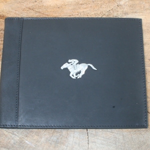 Puede incluir: Álbum de fotos de cuero negro con una imagen en relieve plateada de un jinete montando un caballo.