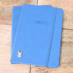 May include: Two blue notebooks with a black elastic band closure. The front cover of the notebook on top has the word "notes" printed in white. A small owl with a graduation cap is attached to the front cover of the notebook on the bottom.