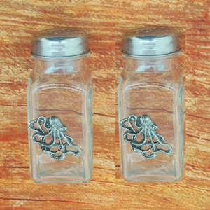Könnte beinhalten: Zwei durchsichtige Salz- und Pfefferstreuer aus Glas mit silbernen Deckeln und einem Tintenfisch-Design auf der Vorderseite.
