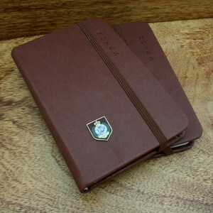 Puede incluir: Dos cuadernos encuadernados en cuero marrón con cierres elásticos. El cuaderno superior tiene una insignia dorada con una corona y las letras "R.C.P.".