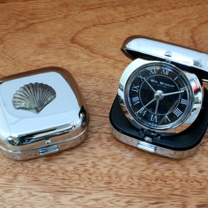 May include: A silver travel alarm clock with a black face and Roman numeral hour markers. The clock is in a silver case with a seashell design on the lid. The clock is made by WM. WIDDOP.