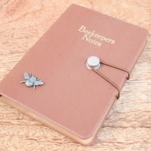 May include: A brown leather-bound notebook with the title "Beekeepers Notes" embossed on the front cover. A silver bee charm is attached to the front cover.