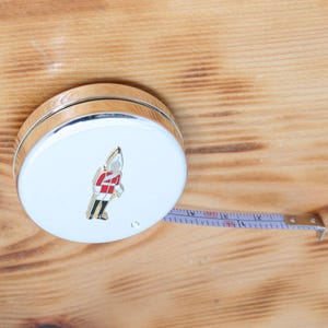 May include: A white and silver tape measure with a decorative soldier emblem. The tape extends from the circular case, showing inch and foot markings. The tape measure is on a wooden surface.