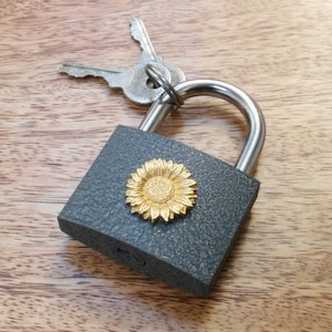 Può includere: Un lucchetto grigio con un design a girasole dorato. Il lucchetto ha una serratura argentata ed è attaccato a due chiavi argentate.