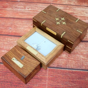 May include: Three wooden boxes with brass accents. The largest box has a decorative brass design on the lid. The middle box has a photo frame with a black and white image of a sailboat. The smallest box has a brass nautical design on the lid.