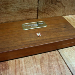 May include: A wooden box with a brass plaque that says "Your Message Engraved Here For Free". The box has a small red and white crest on the front.