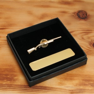 Puede incluir: Un pin de escopeta dorado, colocado en una caja forrada de terciopelo negro con una placa rectangular dorada. El pin es una miniatura detallada de una escopeta. La caja está sobre una superficie de madera.