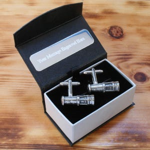 May include: A black gift box with a white interior, containing two silver cufflinks shaped like telescopes. The box has a silver plaque with the text "Your Message Engraved Here".