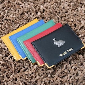 Op de afbeelding: Een stapel kleurrijke reiskaarthouders in geel, blauw, groen, rood en zwart. De zwarte houder heeft een zilveren konijnembleem en de tekst "Travel Card". Elke houder heeft goudkleurige hoekaccenten.