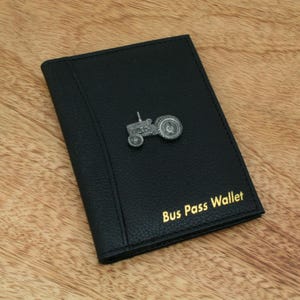 May include: Black bus pass wallet with a silver tractor emblem. The wallet has the words "Bus Pass Wallet" in gold lettering. The wallet is made of a textured material.