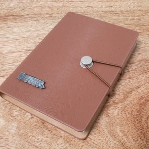 Può includere: Un diario in pelle marrone con un design a forma di locomotiva in metallo sulla copertina. Il diario ha una chiusura a bottone in metallo argentato e un elastico marrone.