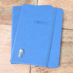 May include: Two blue notebooks with a silver charm attached to the front. The notebook on top has the word "notes" printed on the front.