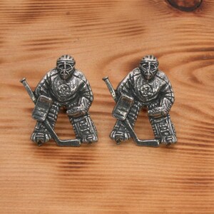 Pu&ograve; includere: Un paio di gemelli argentati con un portiere di hockey in azione. Il portiere indossa un casco e delle protezioni, e tiene un bastone da hockey. I gemelli sono un ottimo regalo per gli appassionati di hockey.