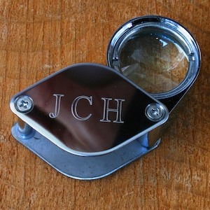 K&ouml;nnte beinhalten: Eine silberne Lupe mit einer runden Linse und einem rechteckigen Griff. Der Griff ist mit den Initialen "JCH" graviert.
