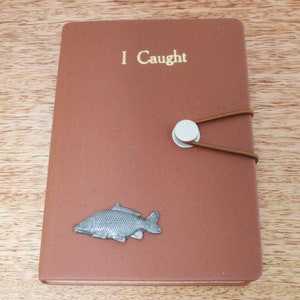 Puede incluir: Un diario encuadernado en cuero marrón con una inscripción dorada que dice "I Caught". Un amuleto de pez plateado está unido a la portada.
