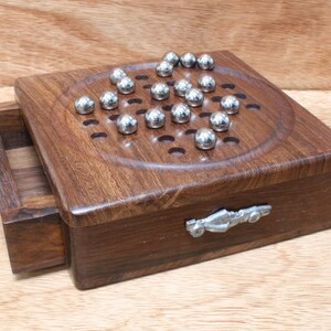 Puede incluir: Una caja de juego de madera con un cajón y una manija de coche de metal. La parte superior de la caja tiene un tablero de juego circular con agujeros y bolas de metal plateadas.