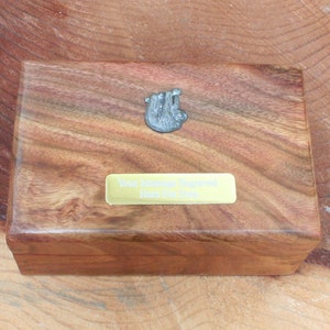 May include: A wooden box with a sloth figurine on the lid. The box has a brass plaque that reads "Your memories preserved here for you".