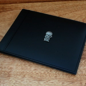 Puede incluir: Una cartera de cuero negro con un pin de metal plateado de una persona sosteniendo un globo terráqueo.