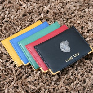 May include: A collection of travel card holders in yellow, blue, green, red, and black. The black holder features a silver emblem and the words "Travel Card" in gold. Each holder has gold corner detailing.