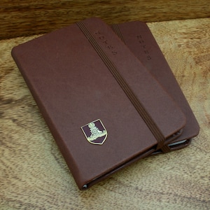 May include: Two brown leather-bound notebooks with elastic closures. The top notebook has a gold emblem with a crown and the words "NOTES" embossed on the cover.