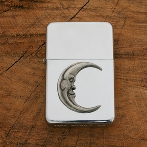 Puede incluir: Encendedor plateado con un dise&ntilde;o de luna creciente. La luna tiene una cara y es de estilo vintage.