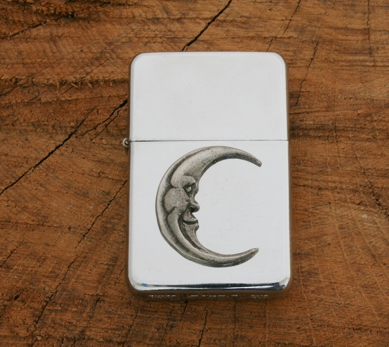Puede incluir: Encendedor plateado con un dise&ntilde;o de luna creciente. La luna tiene una cara y es de estilo vintage.