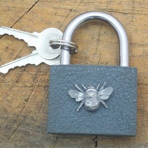 May include: A gray padlock with a silver bee design on the front. The padlock has a silver keyhole and is attached to a silver key ring with two silver keys.