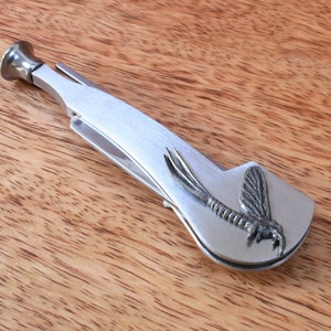 Puede incluir: Una herramienta de metal plateado para pipa con un tampón integrado y un diseño de mosca en el lateral.