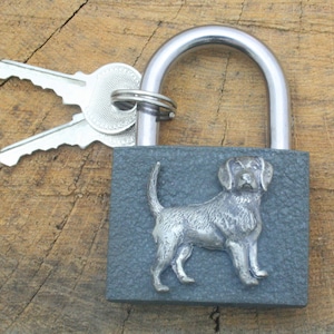 May include: A gray padlock with a silver keyhole and a silver dog silhouette on the front. Two silver keys are attached to the padlock.