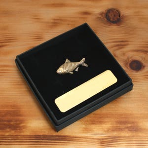 May include: A gold-colored fish-shaped pin resting in a black box with a gold rectangular plate. The pin has detailed scales and fins. The box sits on a wooden surface.