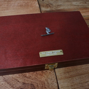 Könnte beinhalten: Eine rechteckige, dunkelrote Holzkiste mit einer silbernen Surferfigur auf dem Deckel. Eine goldfarbene Plakette mit dem Text "Your Message Engraved Here for Free" ist vorne angebracht. Die Kiste hat einen goldfarbenen Verschluss.
