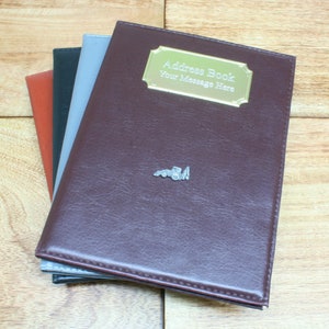May include: A brown leather address book with a gold metal plate that says "Address Book Your Message Here". The book is closed and stacked on top of two other books, one gray and one black.
