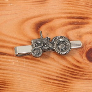 May include: A silver tie clip featuring a detailed miniature tractor with black wheels.