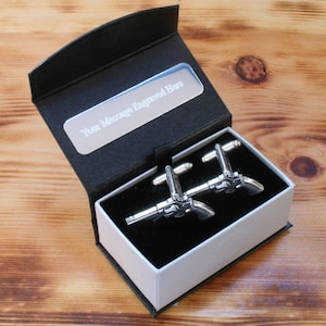May include: A black gift box with a silver inscription that reads "Your Message Engraved Here". The box is open and contains a pair of silver gun-shaped cufflinks.