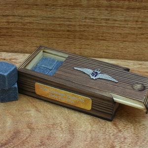 May include: A wooden box with a sliding lid, containing gray stone whiskey chilling cubes. The box has a gold plaque with the text "Your Majesty Enjoyed Here's to the Queen". A silver pin with the letters "RAF" is attached to the box.