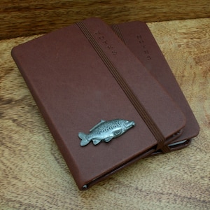 May include: Two brown leather bound notebooks with the word "NOTES" embossed on the front cover. The top notebook has a silver fish charm attached to the elastic closure.