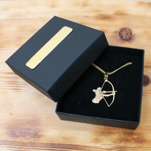 Könnte beinhalten: Eine goldfarbene Halskette mit einem Anhänger in Form eines Bogenschützen, präsentiert in einer offenen schwarzen Geschenkbox mit goldenem Akzent auf dem Deckel. Der Anhänger zeigt einen Bogenschützen mit Pfeil und Bogen. Die Box liegt auf einer Holzoberfläche.