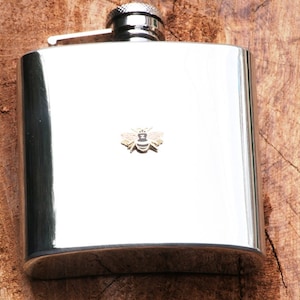 Puede incluir: Un petaca de acero inoxidable con un pequeño emblema de abeja. La petaca tiene una superficie brillante y reflectante y un tapón plateado. El emblema de la abeja es dorado y negro. La petaca está sobre un fondo de madera.