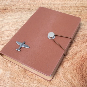 Peut inclure: Un carnet en cuir marron avec un charme d'avion argenté et une fermeture élastique marron. Le carnet est sur une surface en bois.