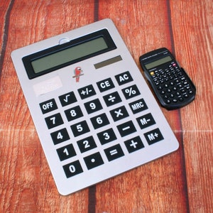May include: A silver calculator with black buttons and a small black calculator are displayed on a wooden surface. The larger calculator has a parrot logo and a digital display. The smaller calculator has many buttons.