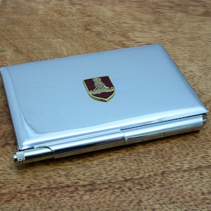 Op de afbeelding: Een zilveren metalen sigarettenkoker met een rood en gouden embleem. Het embleem toont een kroon, een wiel en de tekst "Royal Artillery".